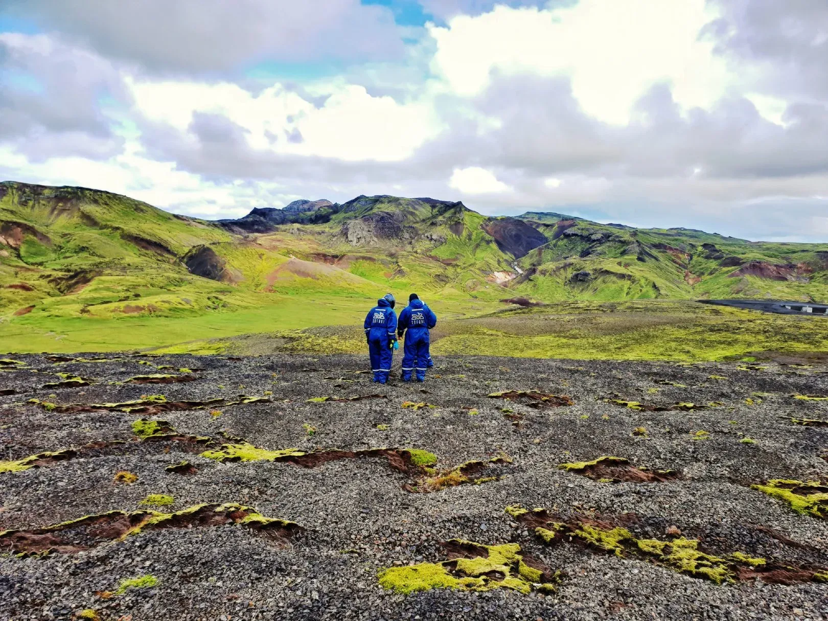 Two people in blue overalls walking across a lava field towards green mountains