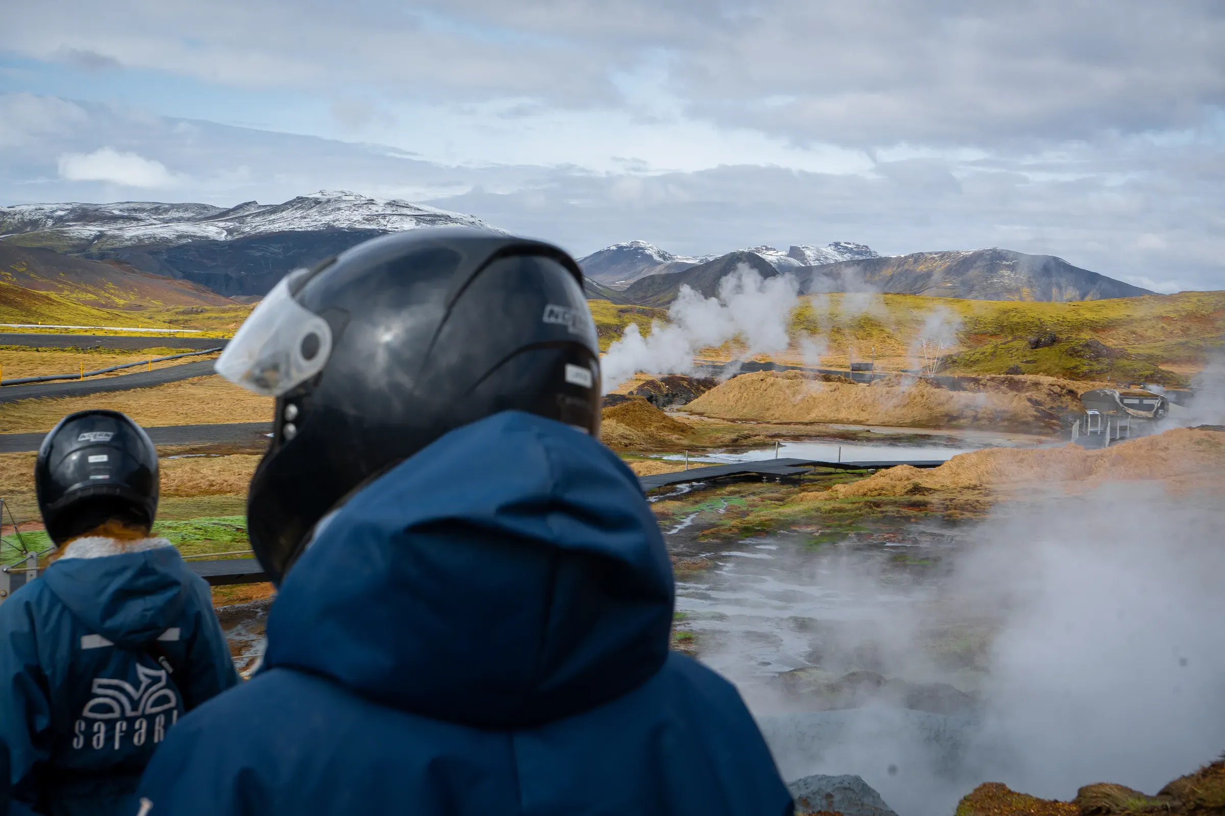 Two riders in helmets looking out over steaming geothermal vents