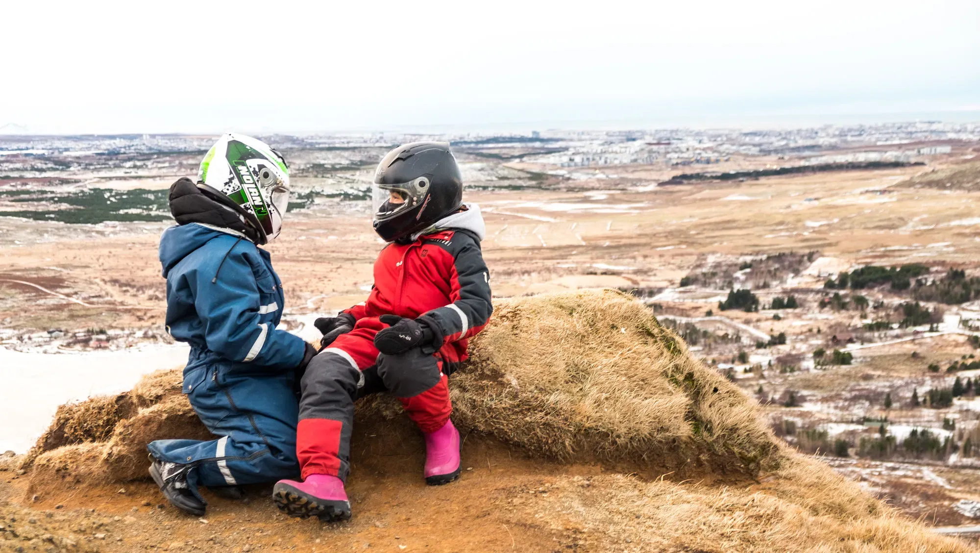 Two children in helmets sitting on a hilltop above Reykjavík