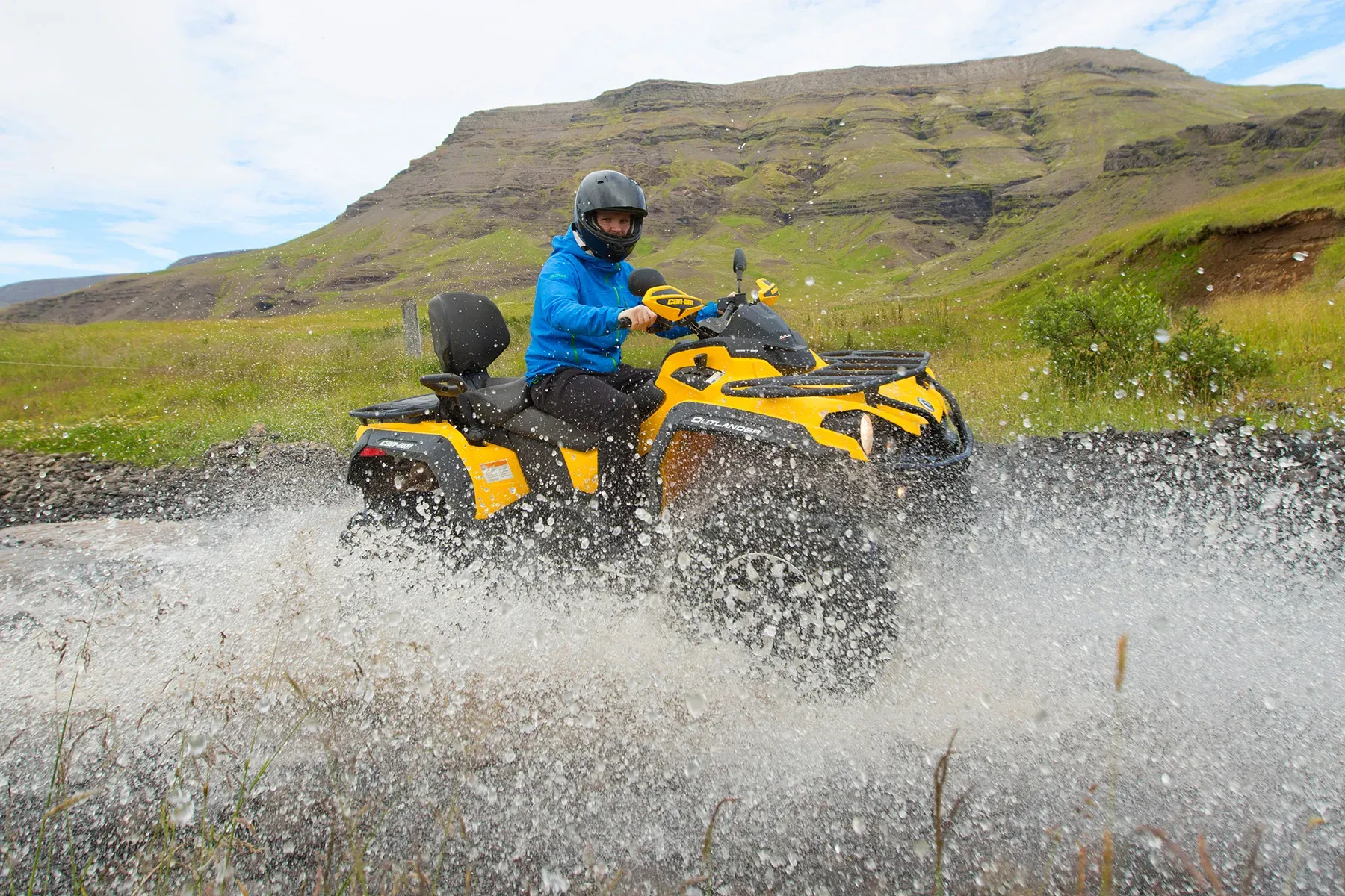Rider in a blue jacket splashing through a river on a yellow ATV