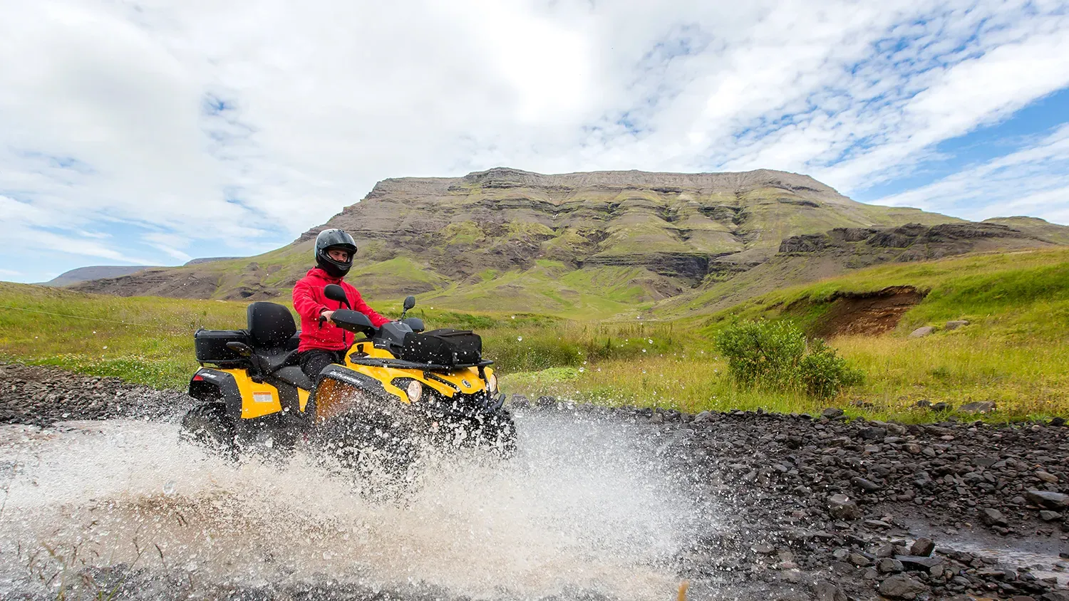 Rider in a red jacket splashing through a river on a yellow ATV